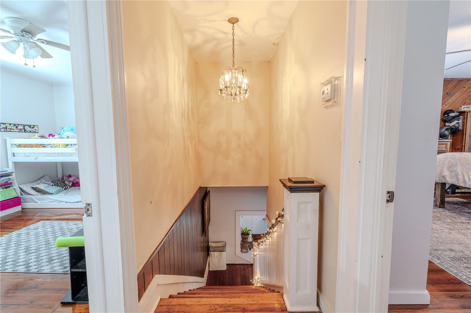This view from the upstairs landing showcases the home’s charming staircase with its classic wood steps, wainscoting detail, and decorative railing. A graceful chandelier casts warm light and elegant shadows, adding character to the space. From the landing, you can access both upper-level bedrooms, highlighting the functional layout and inviting flow of the second floor. The continued presence of original wood flooring enhances the home’s historic charm.