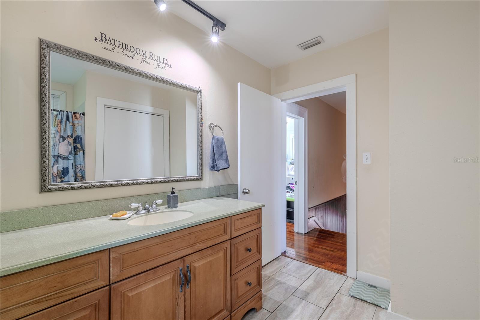 This view from the shower area highlights the spacious layout of the second-floor bathroom, featuring a long Corian countertop with ample vanity storage and a large framed mirror. Tile flooring provides durability, and the doorway opens to the upstairs hallway for easy access to both bedrooms. The generous size and thoughtful design of this bathroom make it a practical and comfortable part of the home.