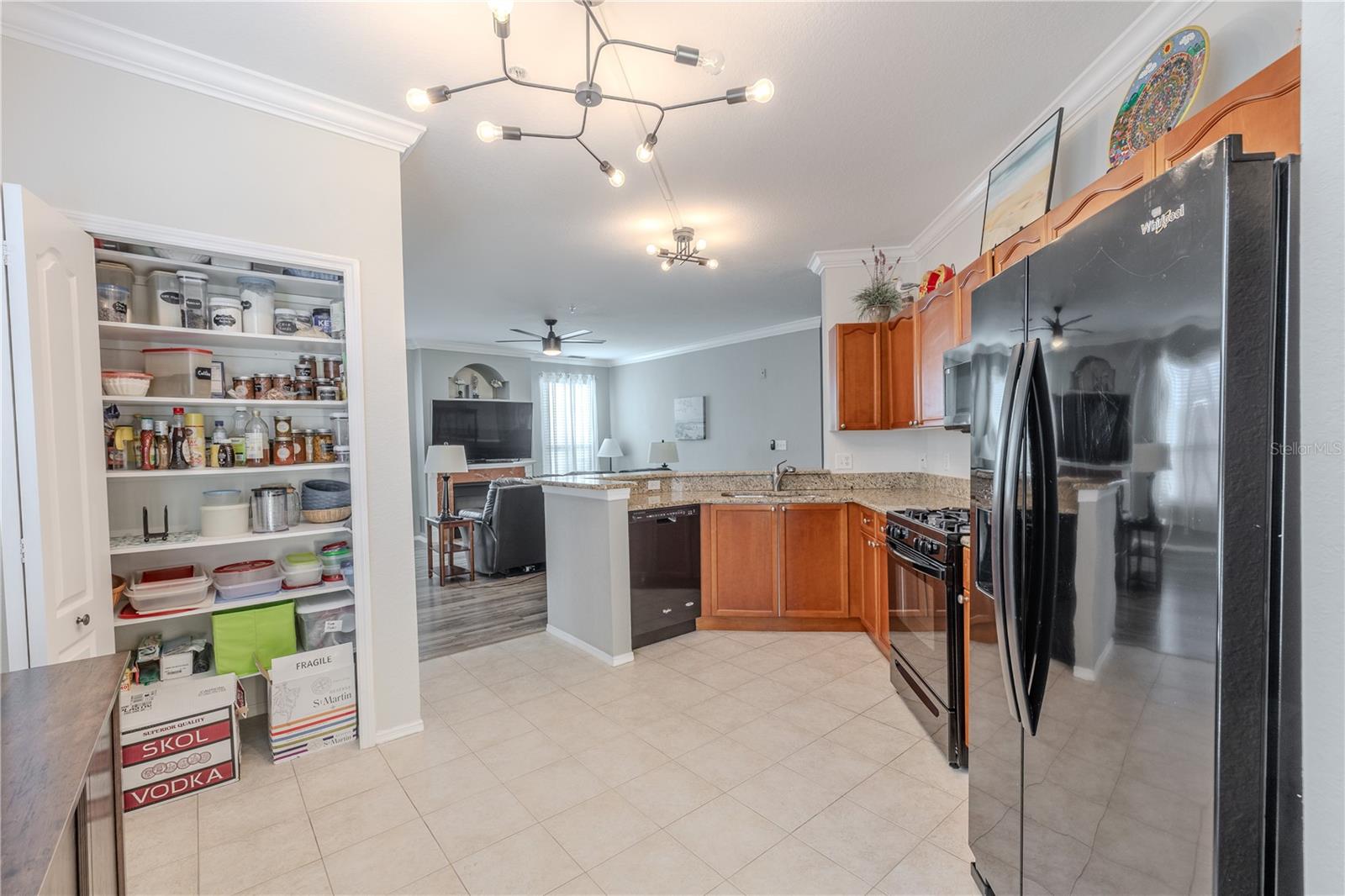 This kitchen view highlights the large pantry with double bi-fold doors, along with granite countertops, stainless appliances, warm wood cabinetry, and an open connection to the living area for seamless everyday living and entertaining.