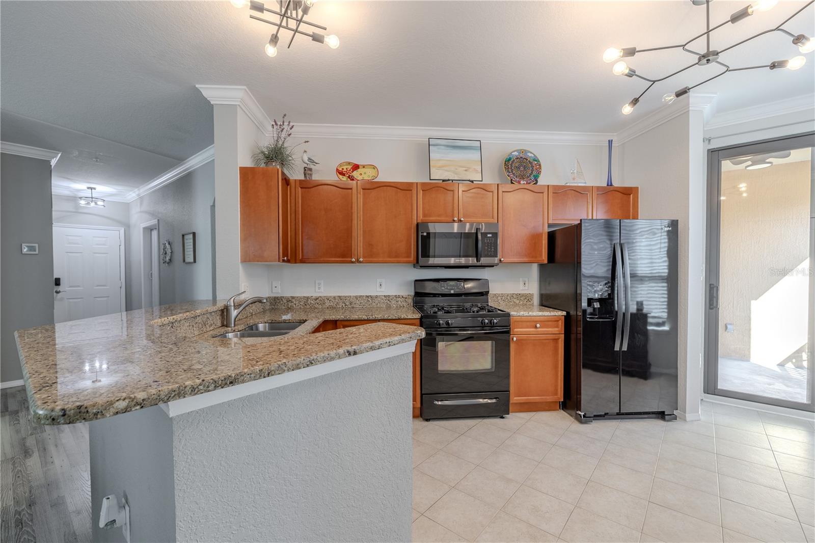 Open kitchen with a spacious granite breakfast bar, warm wood cabinetry, and black appliances, complemented by stylish modern lighting and an easy flow to the living area and screened lanai beyond.