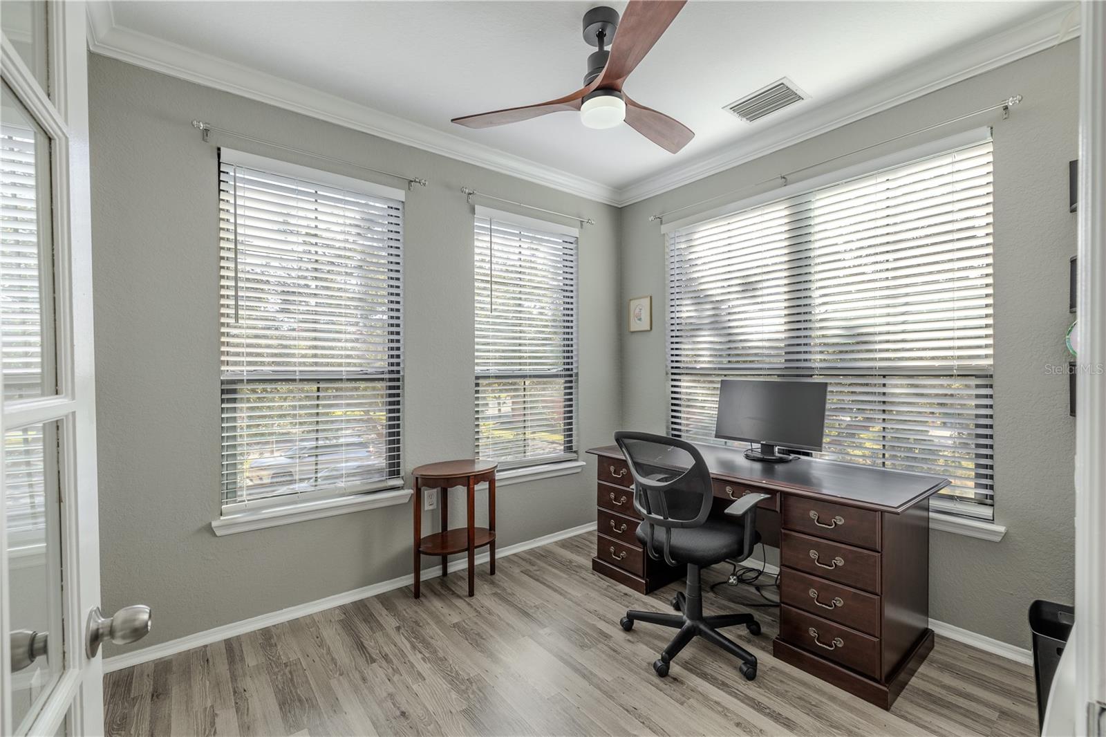 Bright flex room/home office with crown molding, modern ceiling fan, and wraparound windows that fill the space with natural light, complemented by updated flooring.