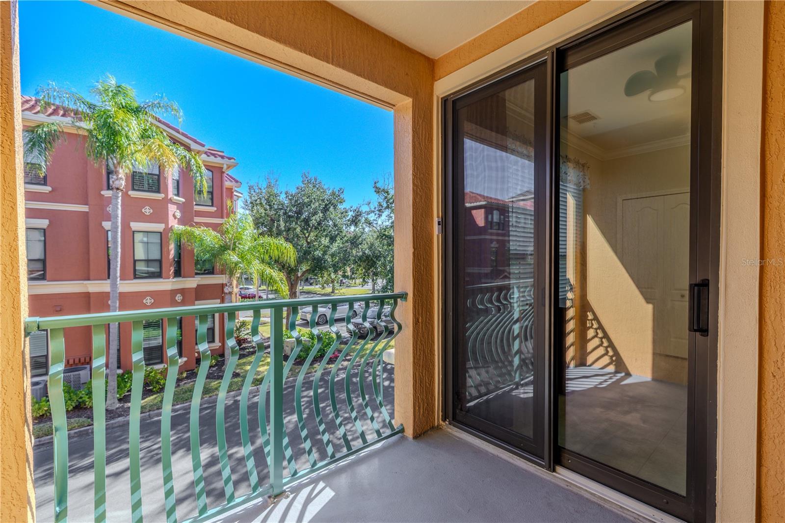 Private covered balcony with sliding glass doors, offering a sunny outdoor space and elevated views of the palm-lined community streetscape.