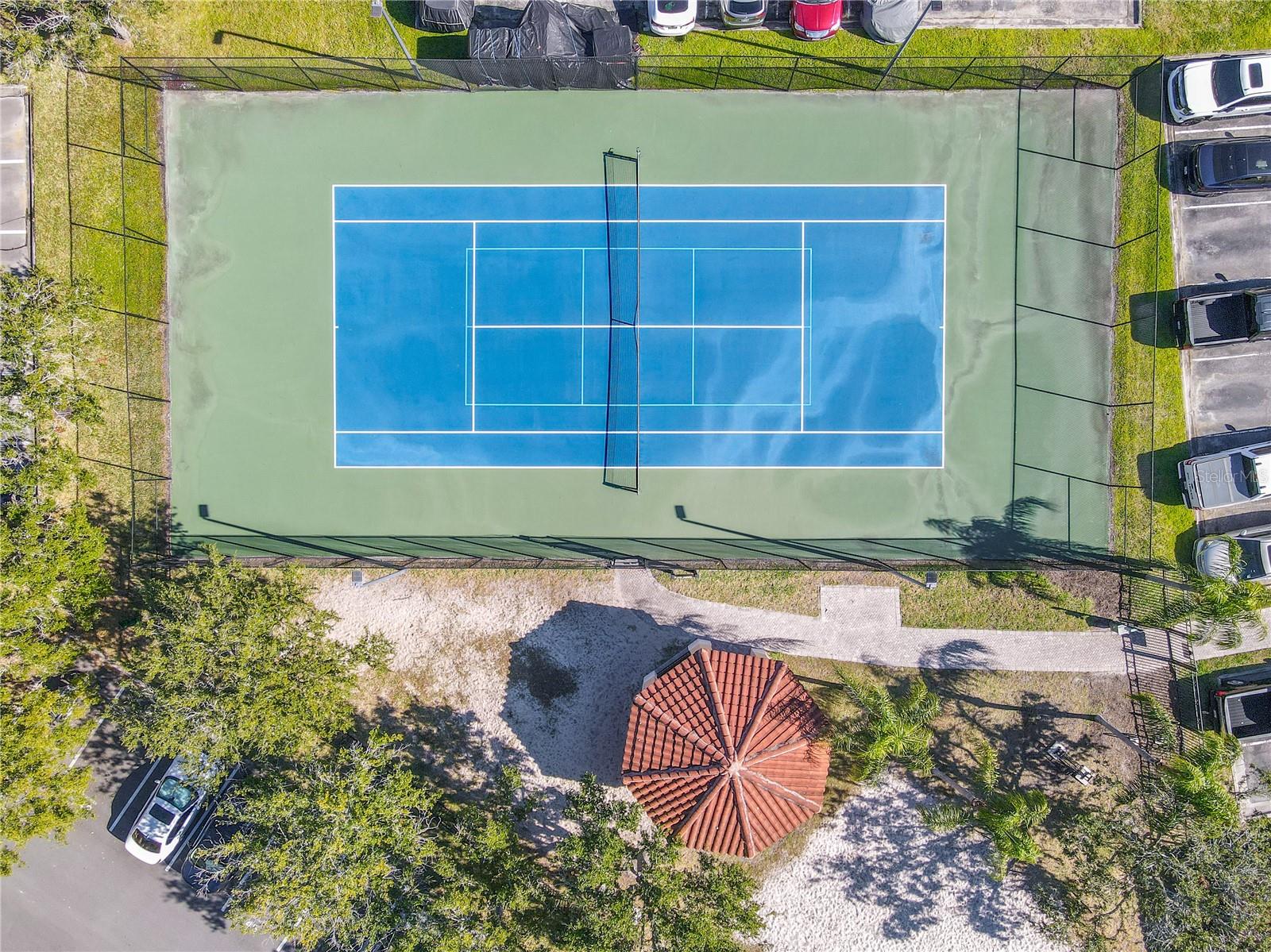 An aerial view highlighting the tranquil community with tennis courtyard with a shaded pavilion, sandy play area, and palm-lined green space, framed by elegant Mediterranean-style residences under clear blue skies.