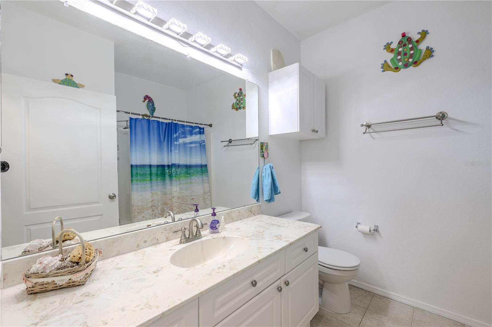 This bright second bathroom features a wide vanity with a solid-surface countertop, ample cabinetry, a full-size tub/shower combination, and cheerful natural light, making it ideal for guests or secondary bedrooms.