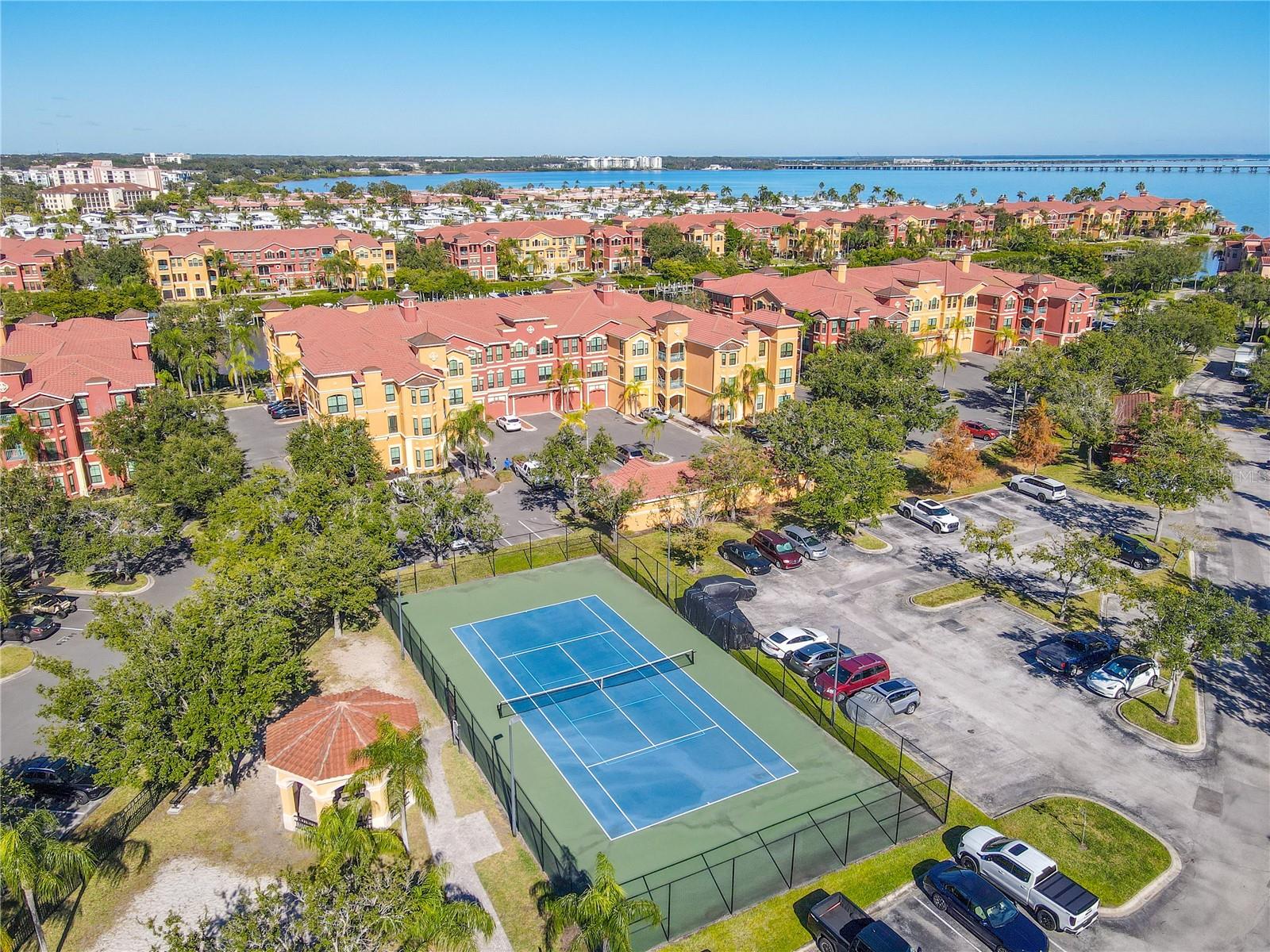 Another aerial view highlighting the tranquil community with tennis courtyard with a shaded pavilion, sandy play area, and palm-lined green space, framed by elegant water view of an the Mediterranean-style residences under clear blue skies.
