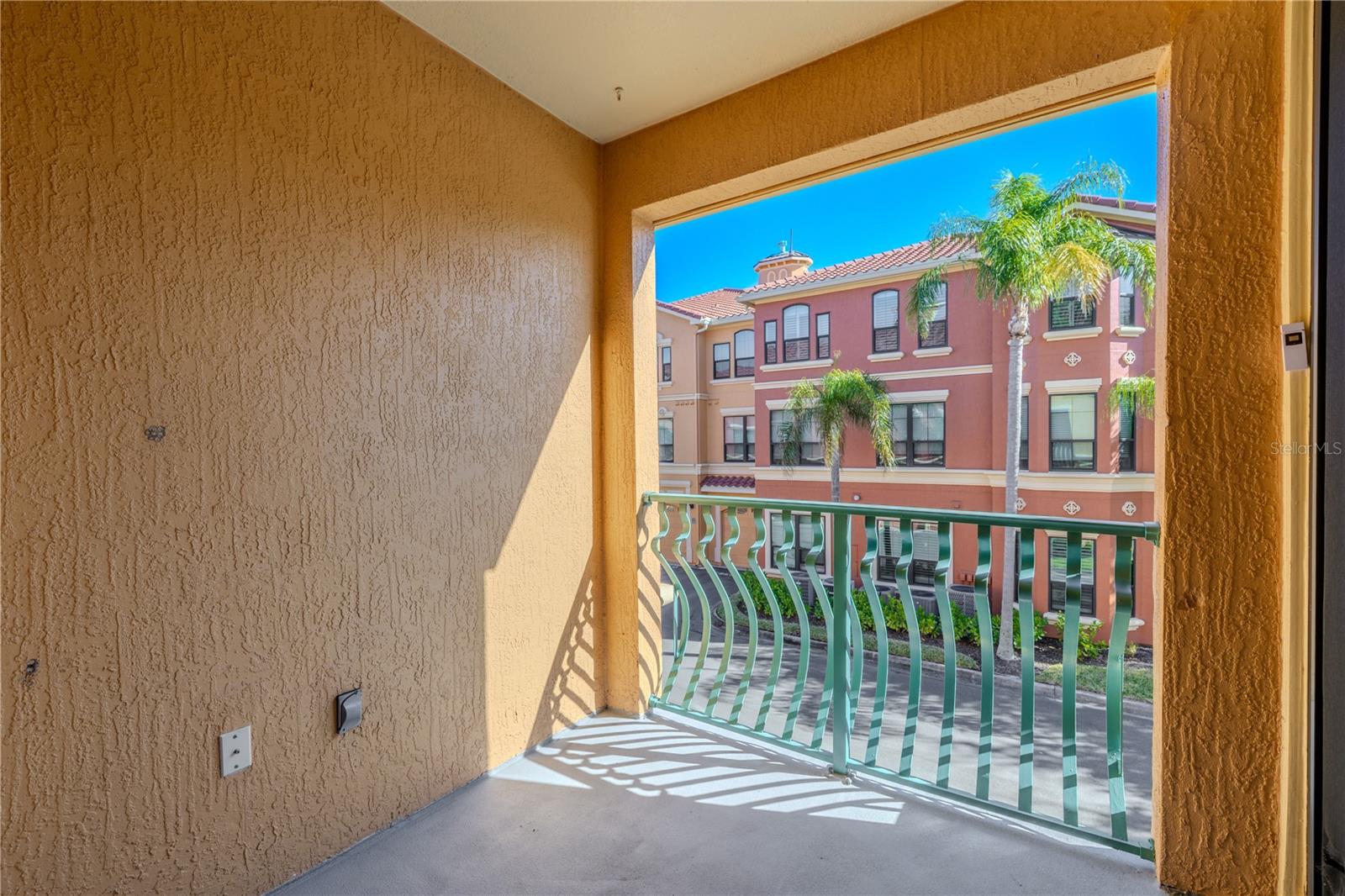 Private covered balcony primary bedroom view with sliding glass doors, offering a sunny outdoor space and elevated views of the palm-lined community streetscape.
