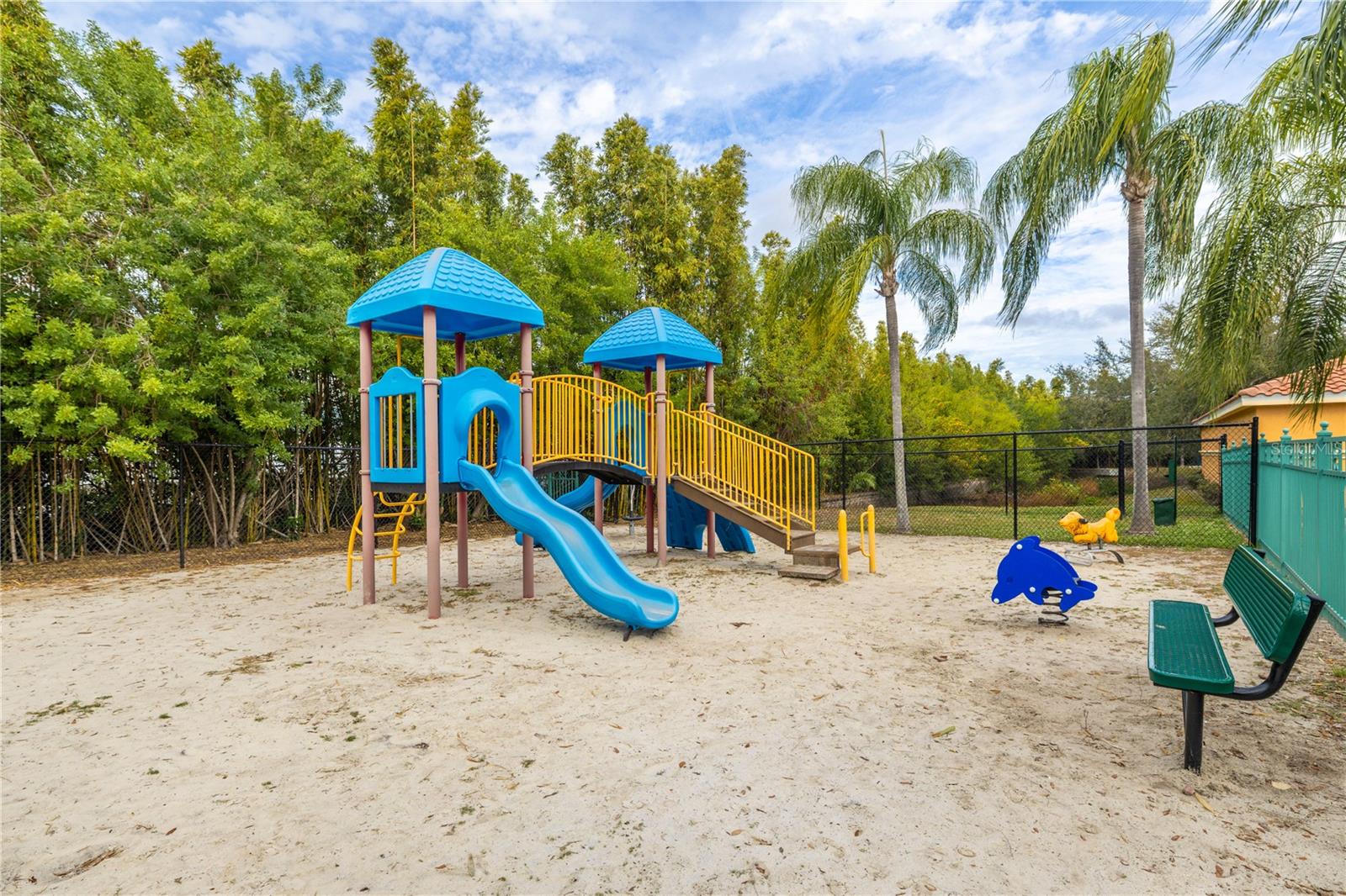 Community playground with a shaded play structure, slides, and seating, set in a sandy play area surrounded by tropical landscaping—perfect for family-friendly fun.