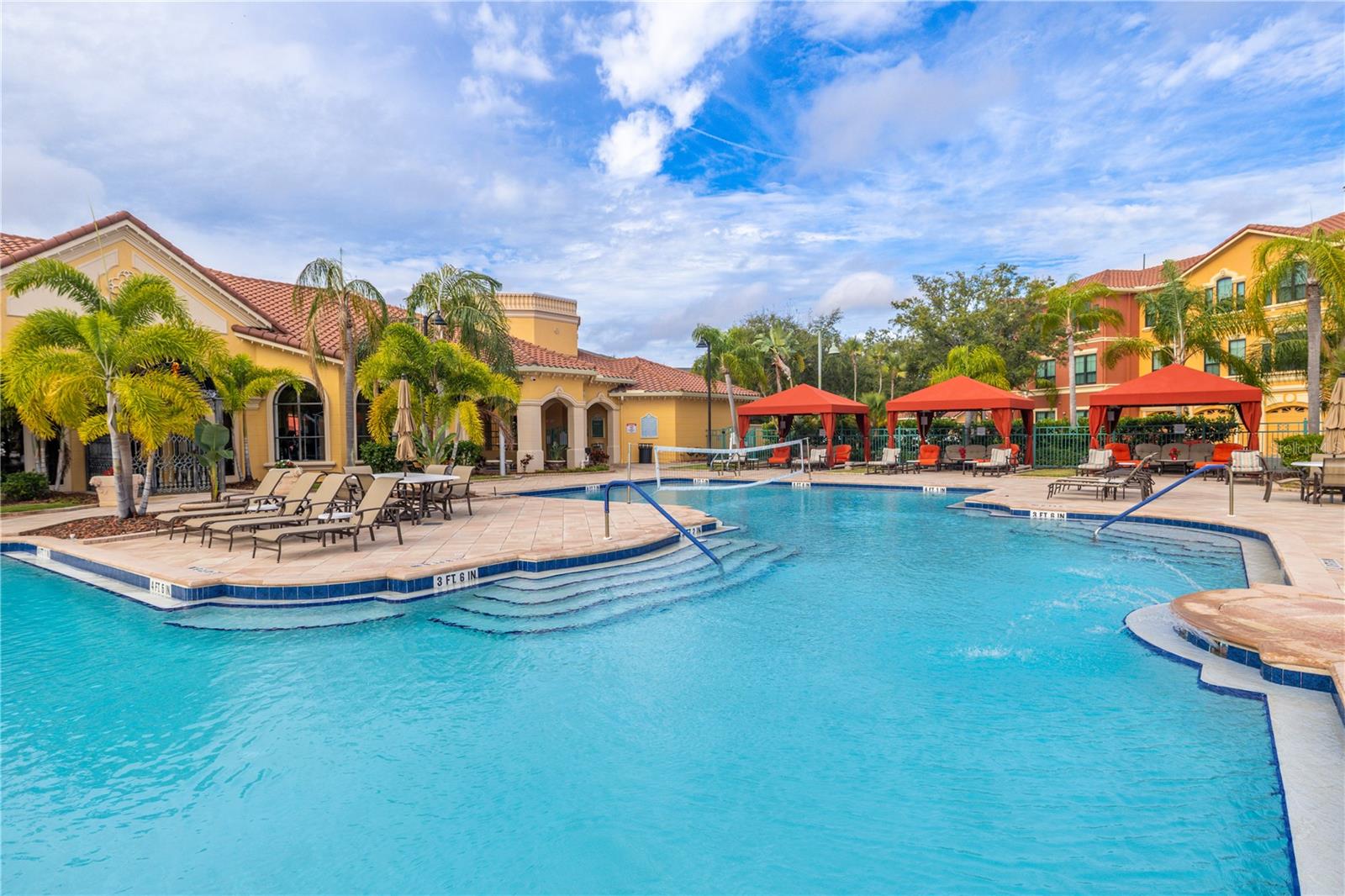 Resort-style clubhouse heated pool with a zero-entry sun shelf, palm-lined deck, lounge seating, and private cabanas—set against beautiful Mediterranean architecture for a true vacation-at-home vibe.
