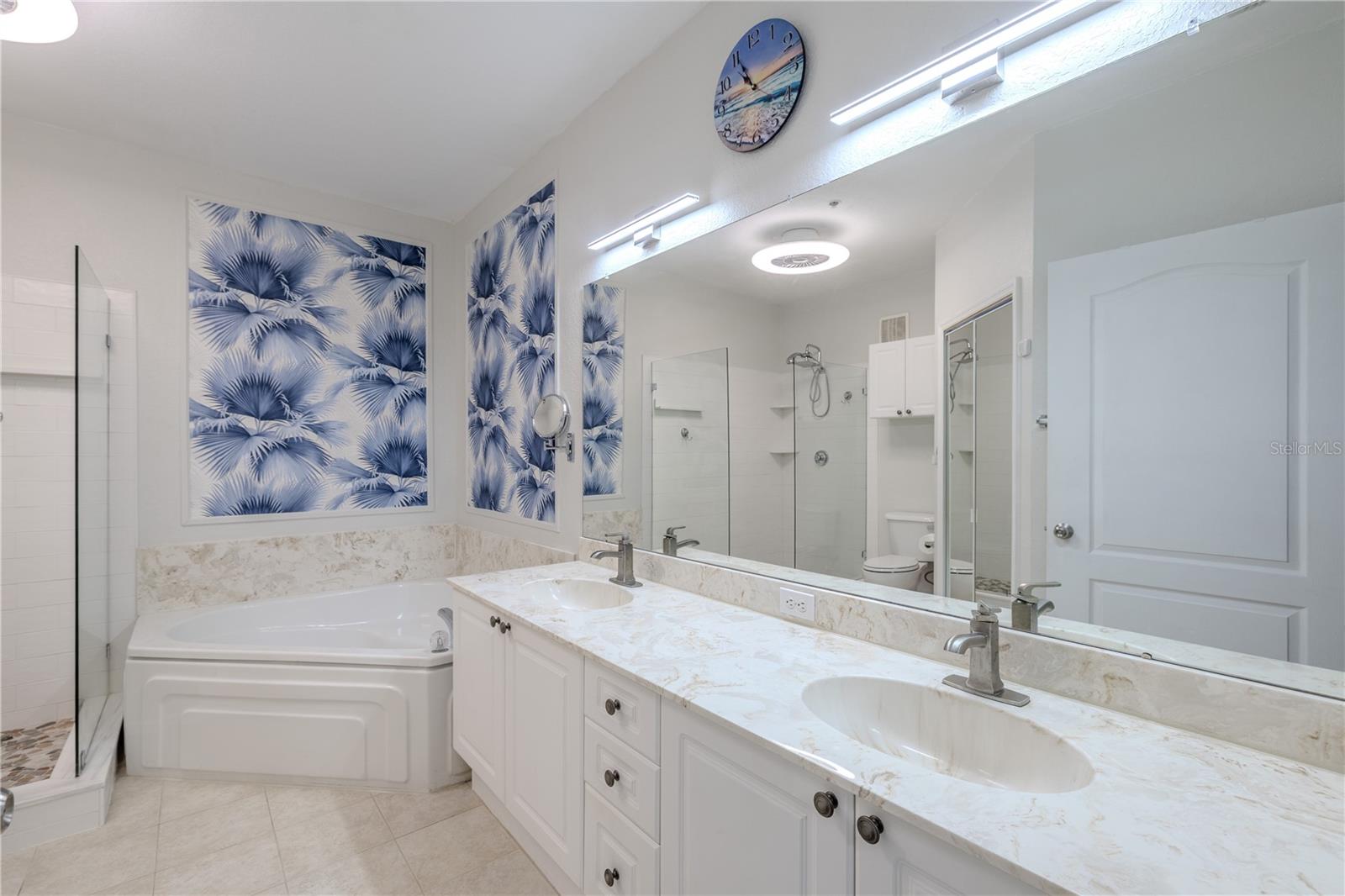 This view highlights the spa-inspired primary bathroom with a glimpse of the glass-enclosed walk-in shower featuring pebble-style flooring, a deep soaking tub with elegant tile surround, and bright, contemporary finishes throughout.