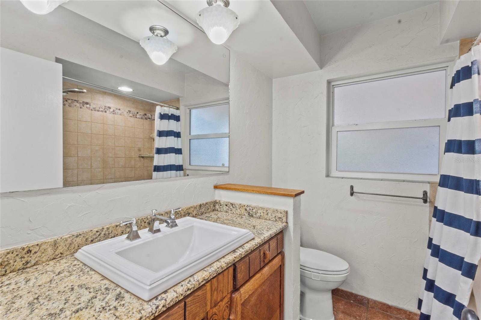 Well-maintained bathroom with granite vanity, framed mirror, tiled shower, and natural light from frosted windows.