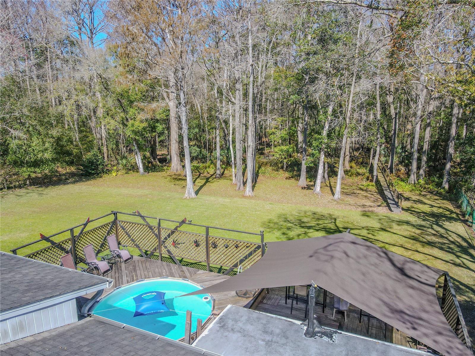 Aerial view highlighting the expansive deck system, above-ground pool, shade structure, and wooded backdrop beyond the yard.