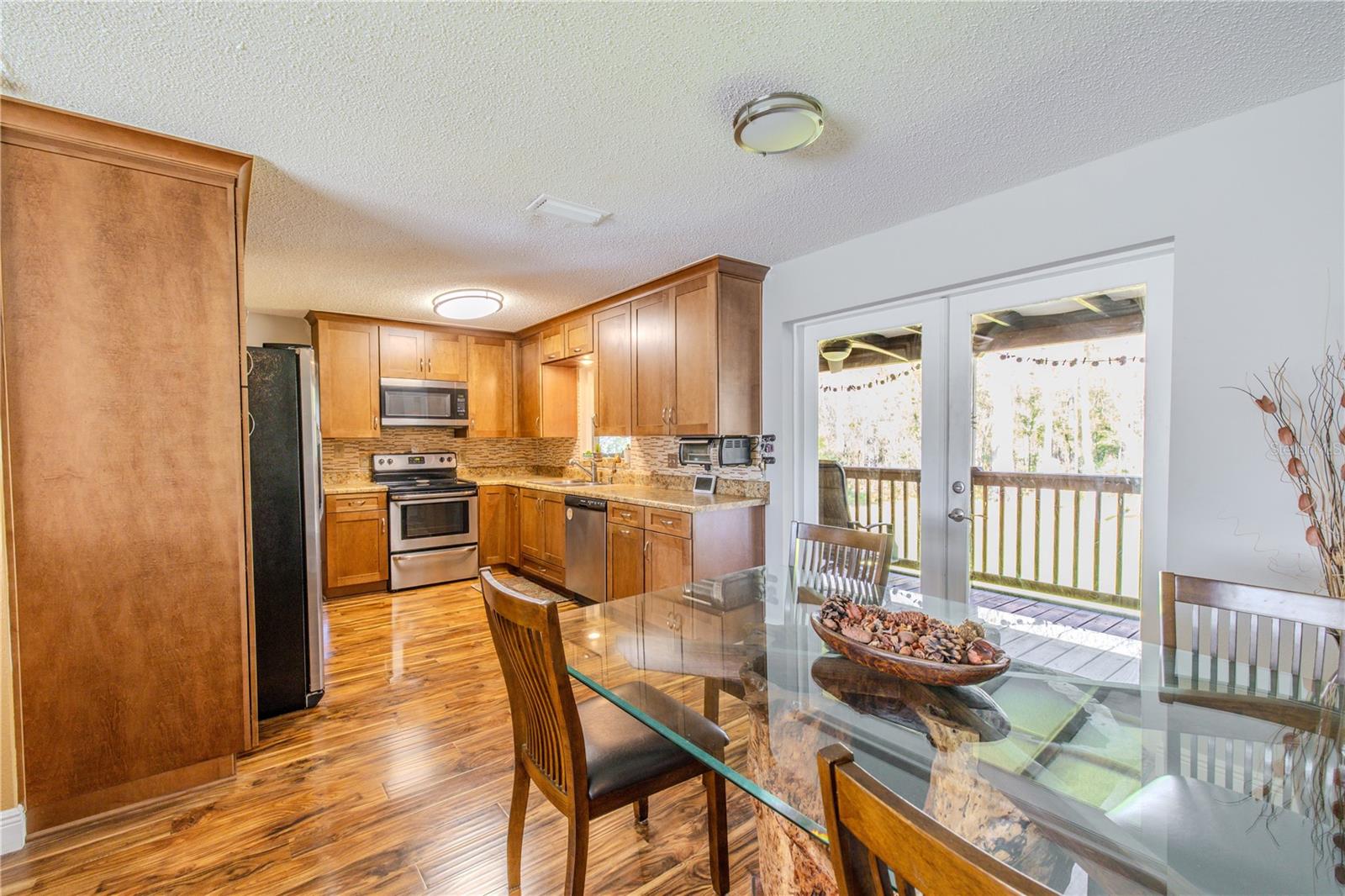 Warm, well-appointed kitchen featuring solid wood cabinetry with crown molding, granite countertops, and a coordinating mosaic tile backsplash. Stainless steel appliances include a smooth-top range, built-in microwave, dishwasher, and side-by-side refrigerator. The laminate flooring flows seamlessly into the dining area, where glass doors invite natural light and open to the covered deck beyond.