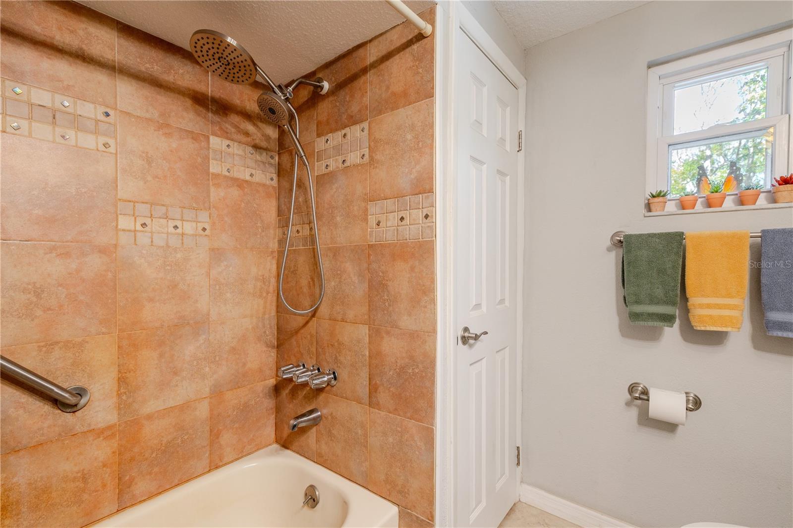 Second bathroom tub/shower combination with tiled surround, decorative accent inlay, grab bar, and rainfall-style showerhead.