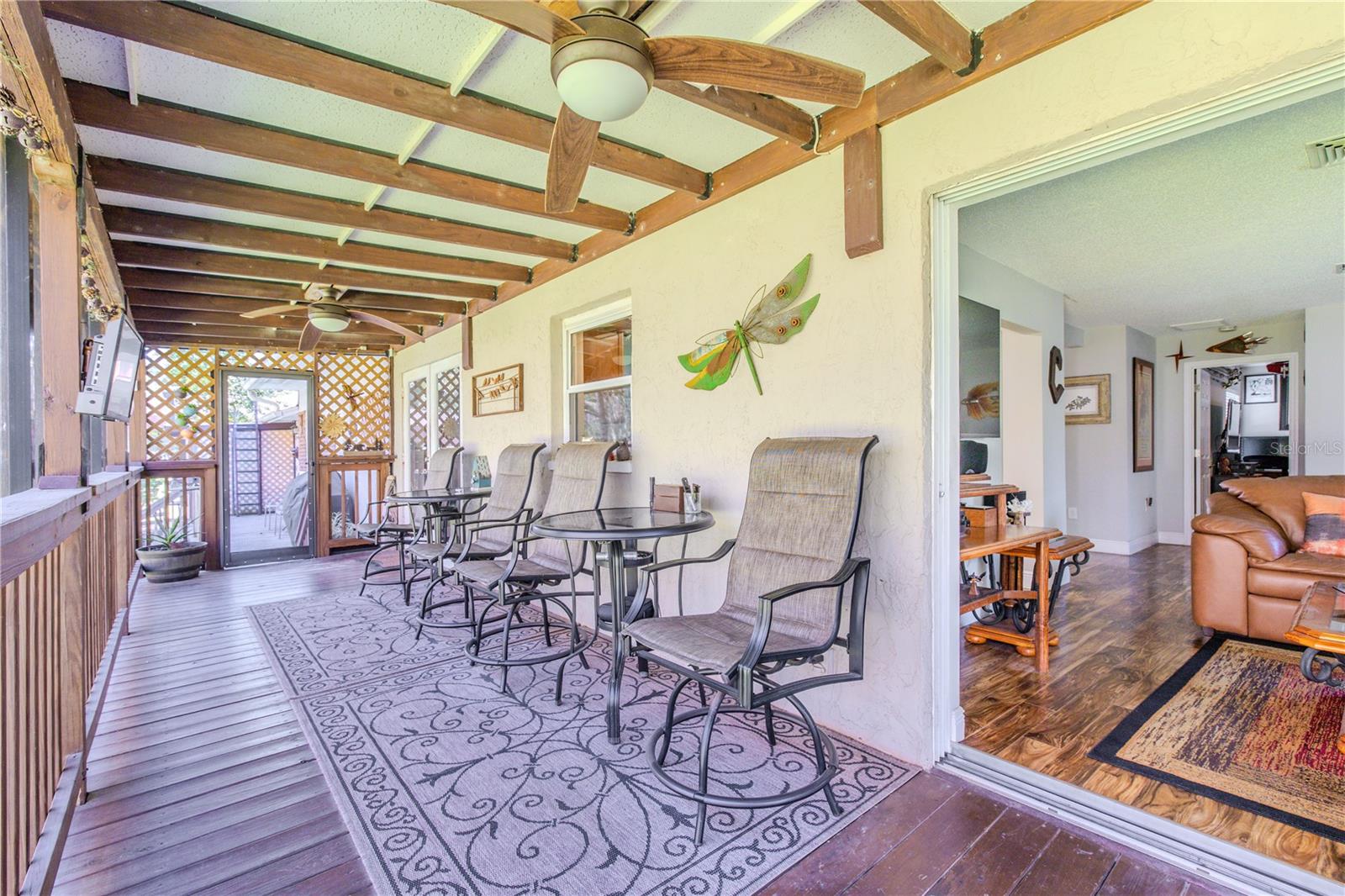 This expansive covered deck stretches the length of the home, thoughtfully arranged with multiple options for seating areas. A pergola-style ceiling with ceiling fans provides shade and comfort, while lattice accents and wood railings frame peaceful views. The layout allows easy flow between the interior living space and the outdoors.