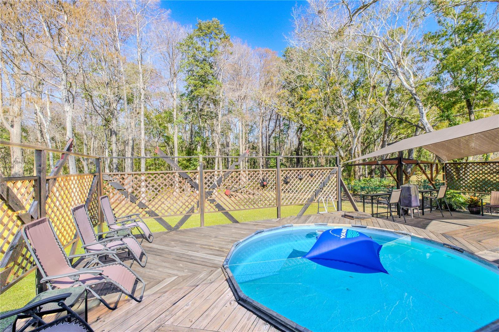 An above-ground pool is integrated into a spacious wood deck, framed by lattice fencing for privacy and safety. The setting is open yet secluded, with peaceful views of surrounding trees and natural landscaping.