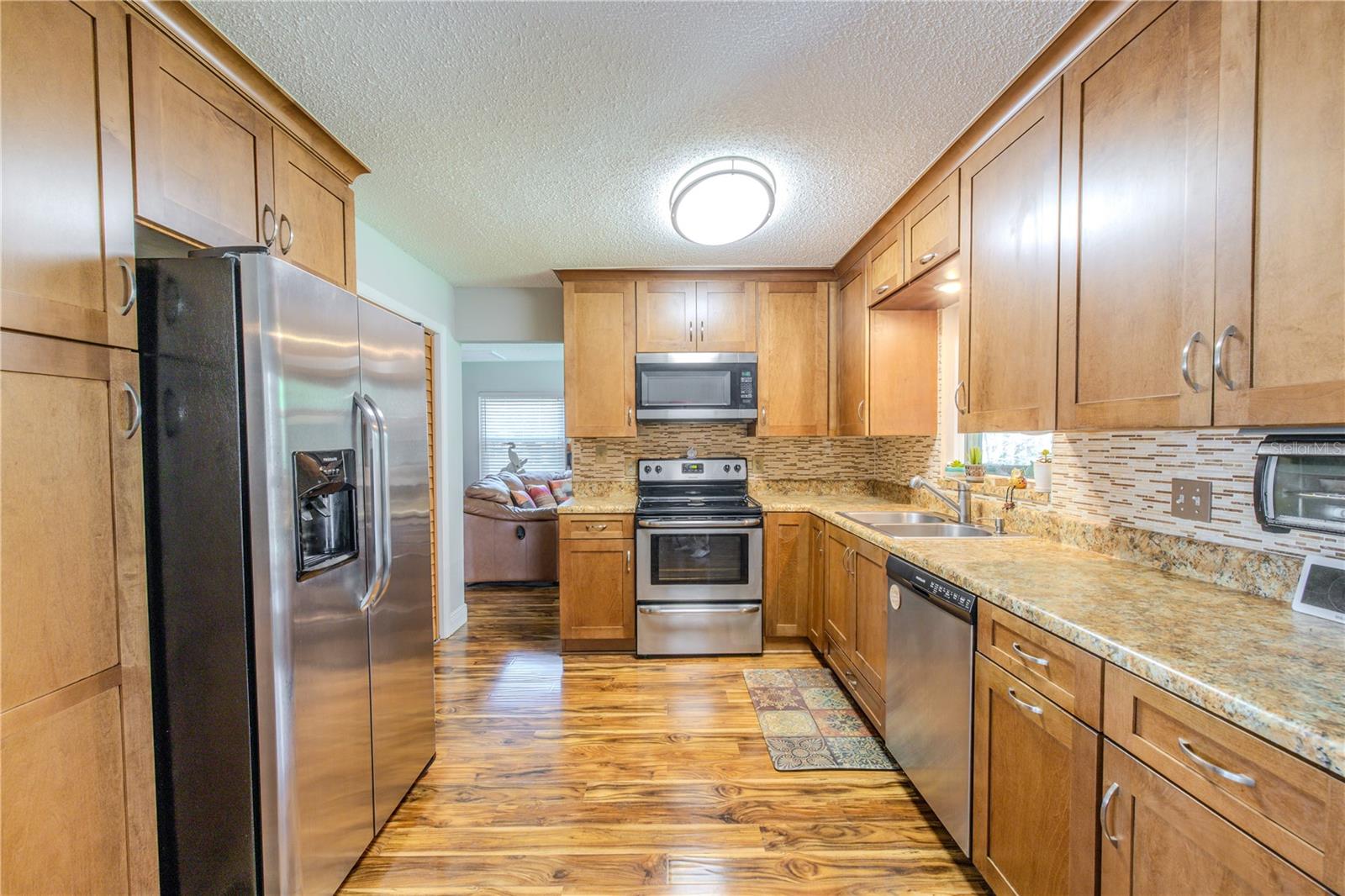 Well-designed kitchen featuring warm wood cabinetry, granite countertops, mosaic tile backsplash, and laminate flooring, with stainless steel refrigerator, range, microwave, and dishwasher arranged in an efficient galley-style layout.