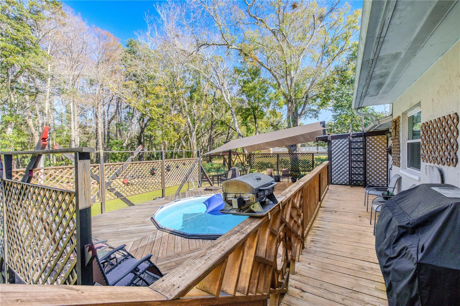 A multi-level wood deck creates a private outdoor retreat, surrounded by lattice accents and mature trees. The layout offers defined zones for recreation and relaxation, with railings and open sightlines overlooking the yard and wooded backdrop.