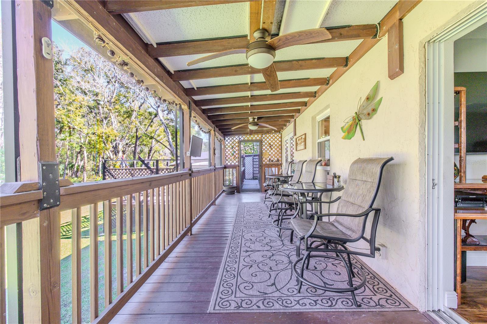 A long, covered deck offers ample space for seating and entertaining. Ceiling fans and a pergola-style roof provide year-round comfort, while open railings and surrounding greenery create a relaxed, private setting. Multiple access points to the interior enhance functionality and flow.