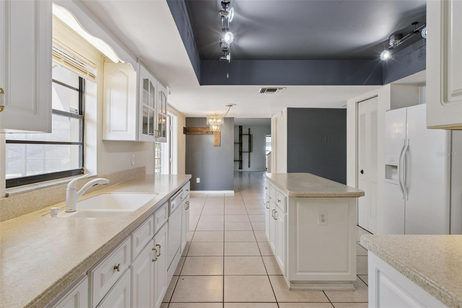 Kitchen view looking past the center island toward the adjacent living areas. White cabinetry and light countertops line both sides of the kitchen, with the island centered in the walkway. A refrigerator is on the right, and tiled flooring continues into the surrounding rooms. Track lighting is installed within a recessed ceiling feature above the kitchen.