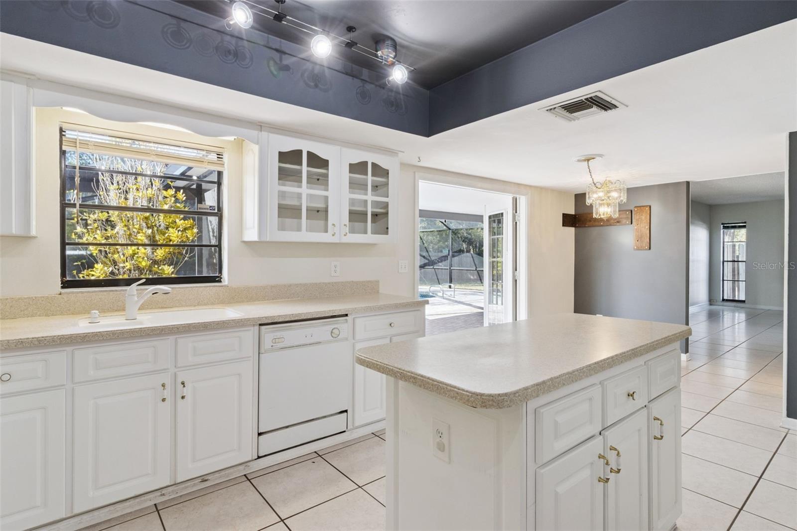 View from the kitchen showing the sink beneath a window on the left and the center island in the foreground. White cabinets with glass-front upper doors are visible. Beyond the island, the space opens toward the breakfast nook area, with natural light entering through glass doors. Tile flooring runs throughout the kitchen and into the adjoining space.