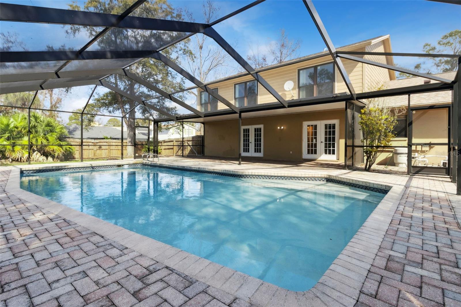 Wide view of the pool and screened enclosure showing the rear exterior of the home. Multiple sets of glass doors open from the house onto the pool deck. The pool sits centrally within the enclosure, surrounded by brick pavers, with the screen roof and support framing visible overhead.