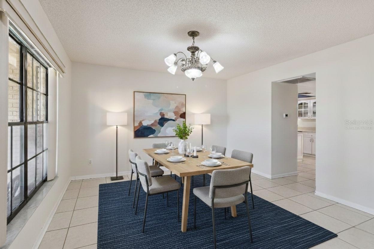 This photo is virtually staged. Formal dining room featuring tile flooring, neutral wall colors, and a centrally located ceiling-mounted light fixture. A large window provides natural light. The space is virtually staged with a rectangular dining table, chairs, area rug, and accent lighting to illustrate possible furniture placement.