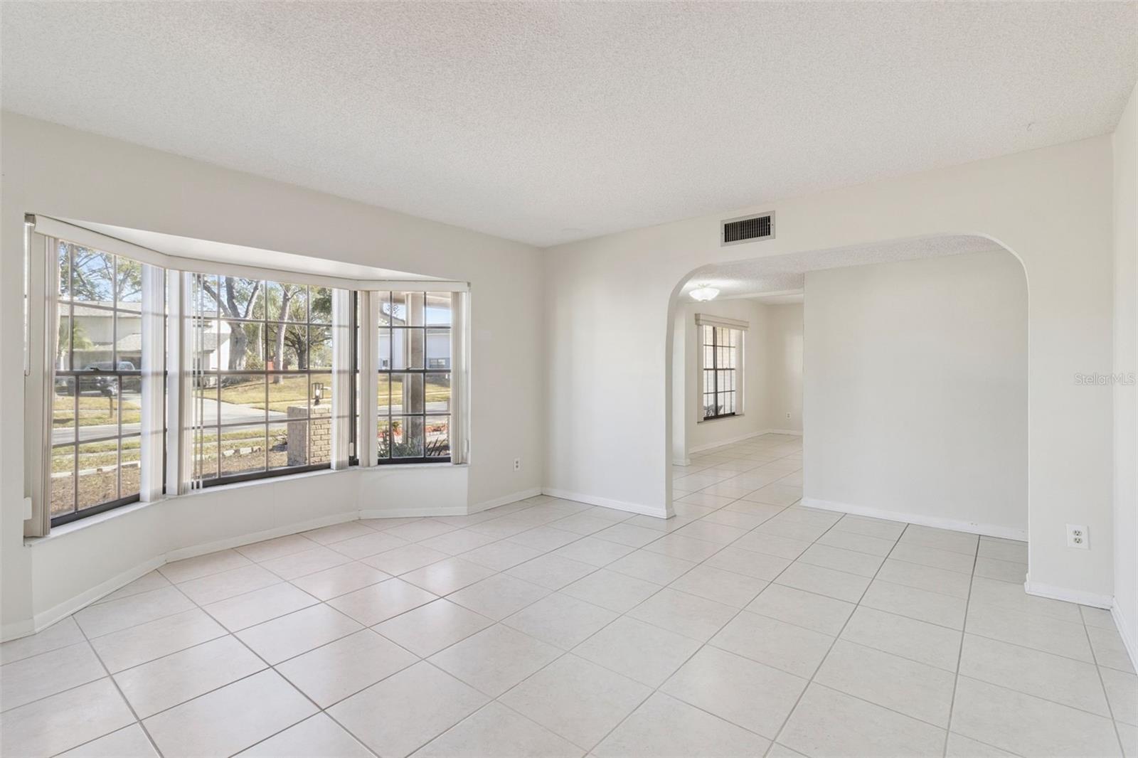 Alternate angle of the formal living room showing the bay window from the interior and an arched opening leading to the formal dining room. Tile flooring continues throughout the space.
