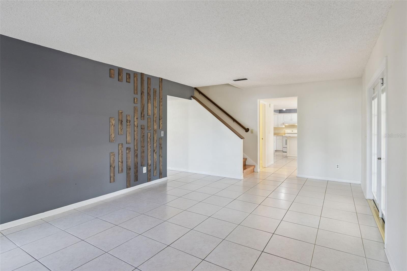 Alternate view of the family room showing the gray accent wall with wood accents, tiled flooring, and an open layout connecting to adjacent living spaces. A staircase with wood handrail is visible along one wall.