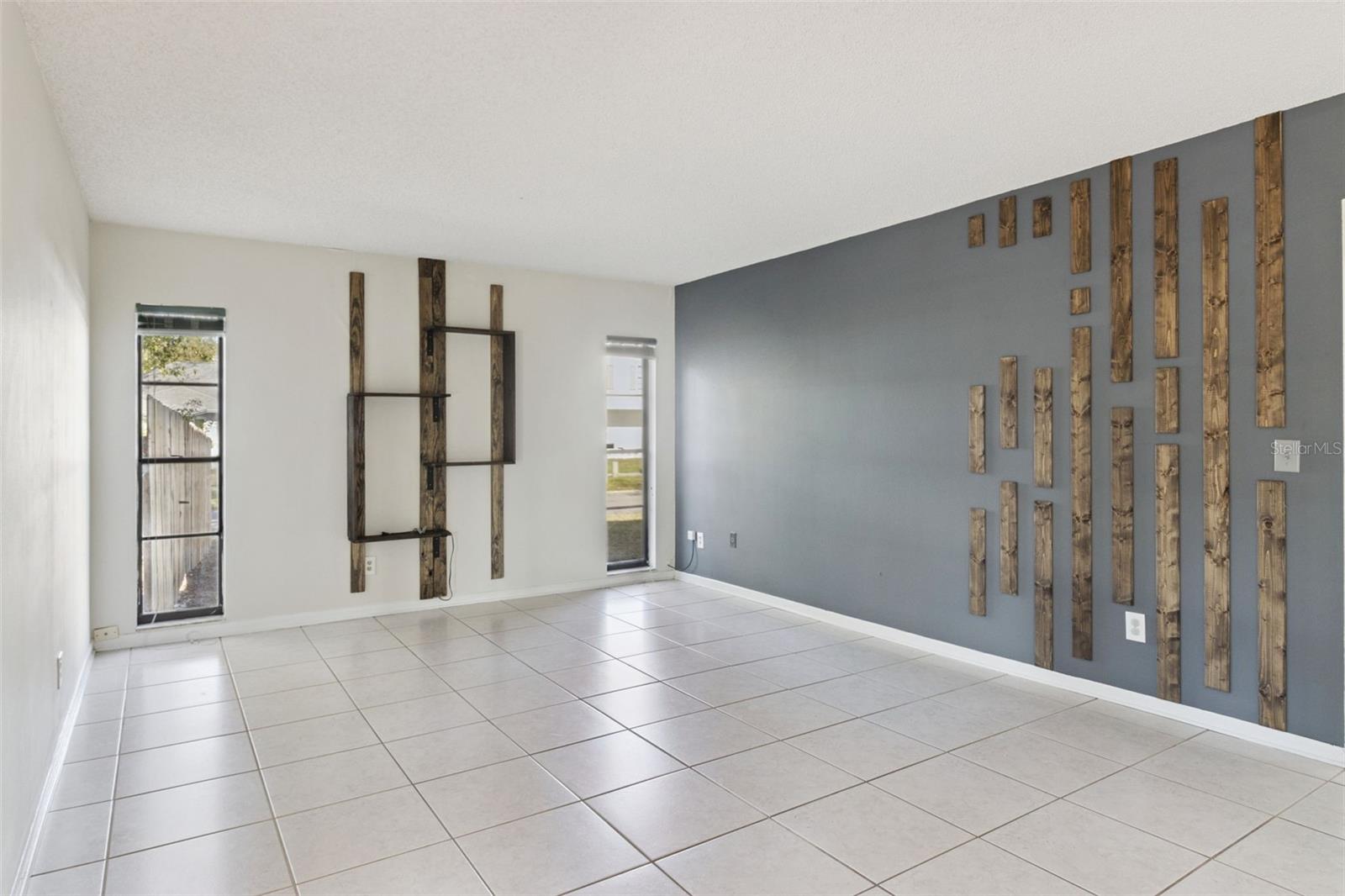 Spacious family room with light-colored tile flooring and neutral walls. A gray accent wall features decorative vertical wood accents. The room includes multiple windows that allow natural light and provide views of the yard.