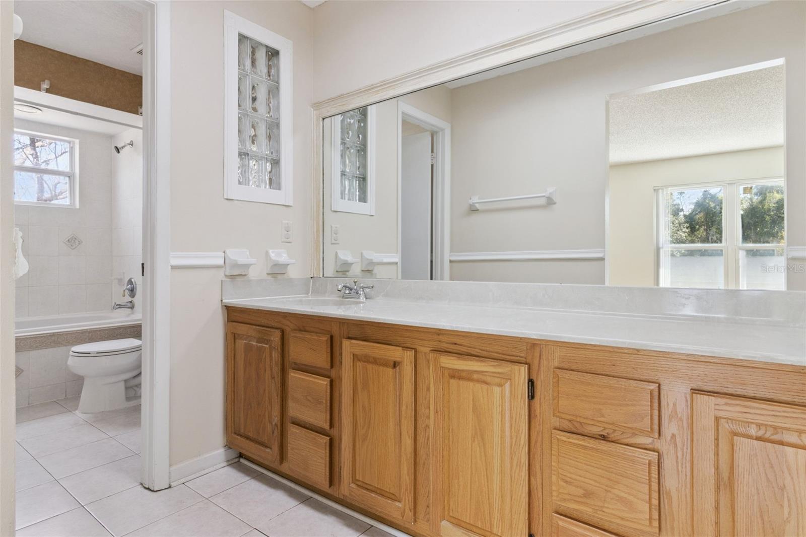 Primary bathroom vanity area with an extended countertop, wood cabinetry, and a large wall-mounted mirror. Tile flooring and neutral wall tones create a bright, open feel.