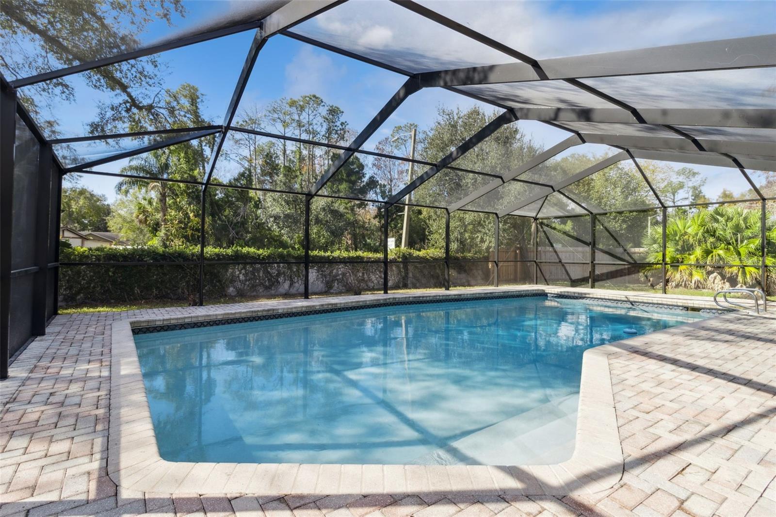 Angled view of the in-ground pool inside the screened enclosure. The pool is bordered by brick pavers on all sides, with a metal pool ladder visible at one end. The screen structure frames views of surrounding landscaping and neighboring properties beyond the enclosure.