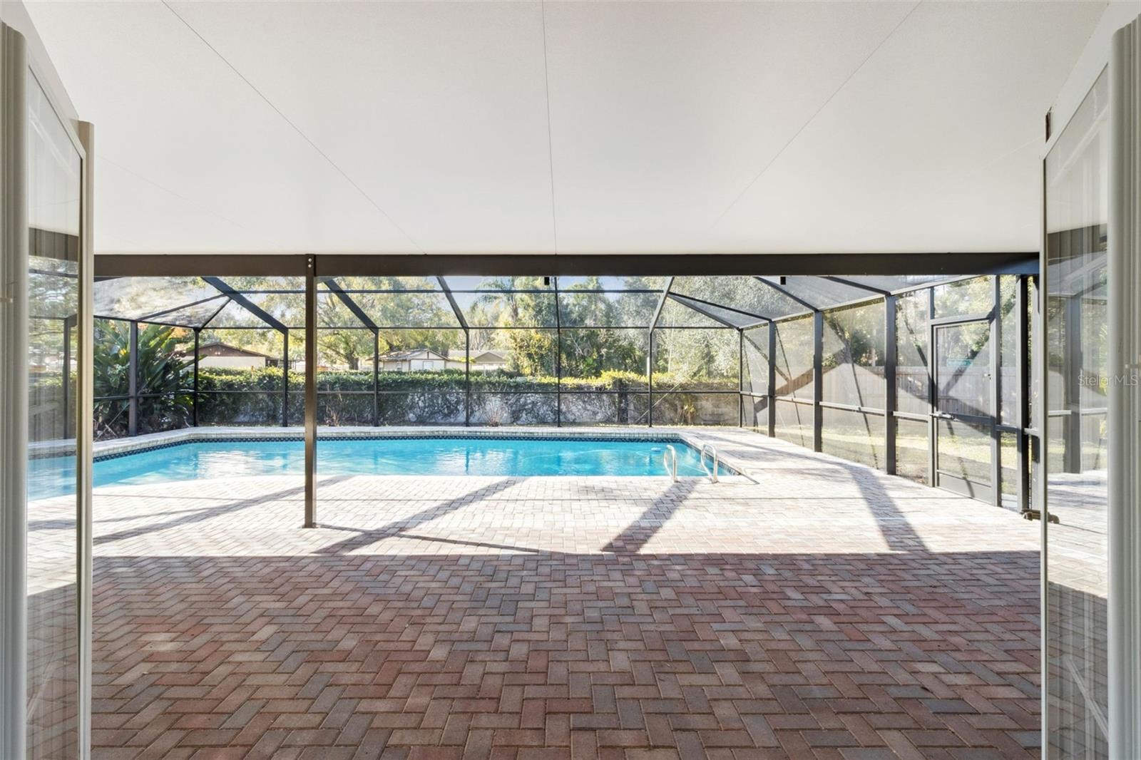 View from the covered portion of the screened enclosure looking out toward the pool. Brick pavers extend across the deck area, leading to a rectangular in-ground pool with light-colored coping. The screen enclosure structure surrounds the space, with trees and fencing visible beyond the screen panels.