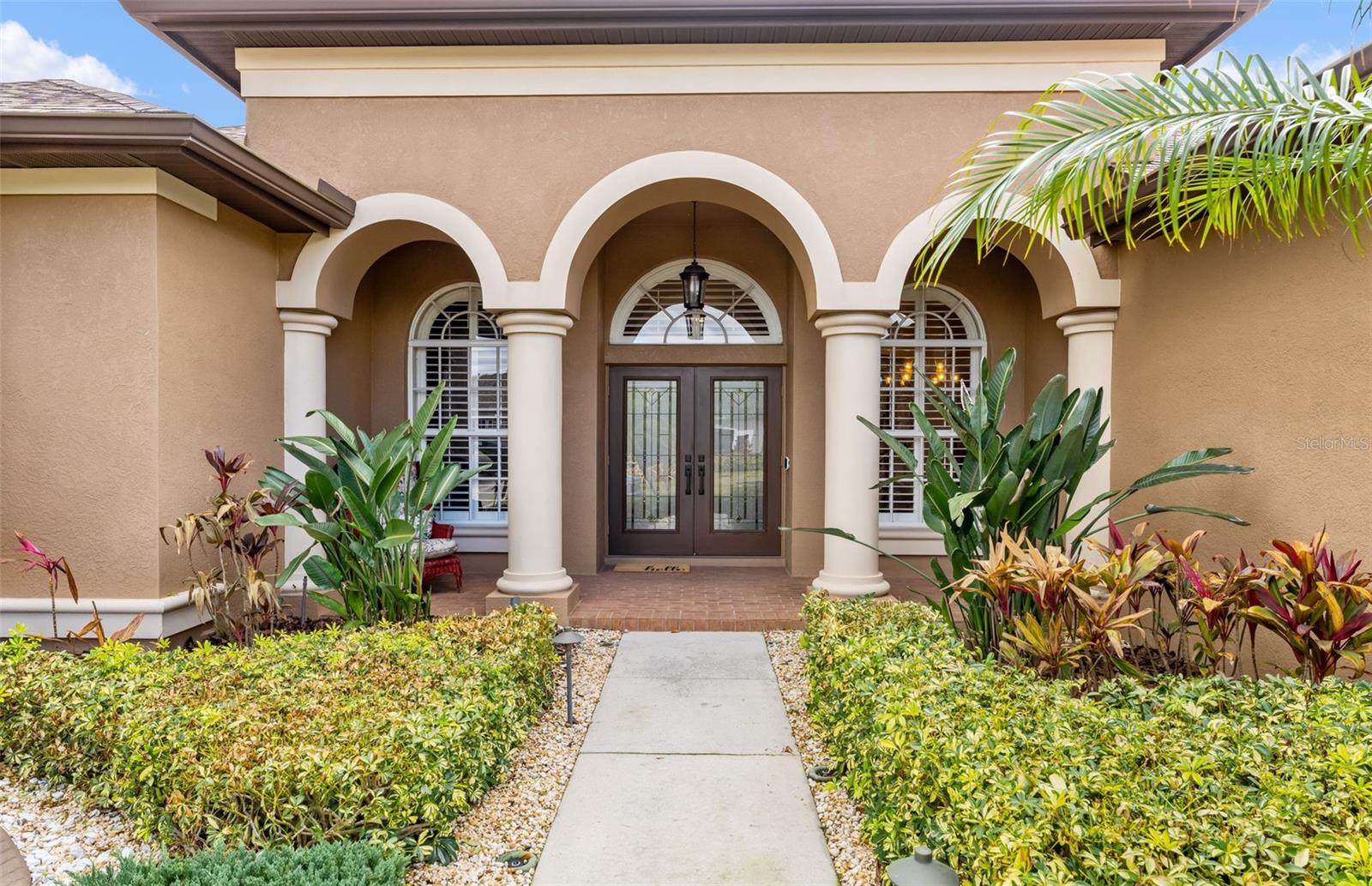 A grand arched entryway with stately columns and double glass doors sets an elegant tone, framed by mature landscaping and a charming brick walkway.