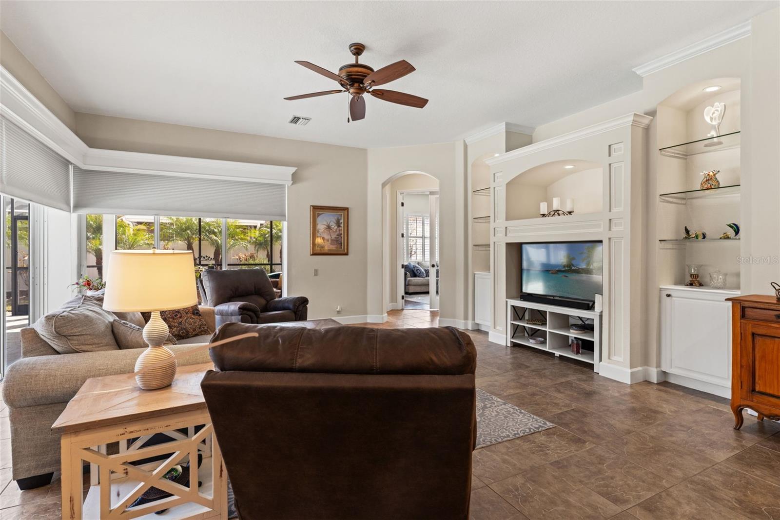 The comfortable family room features a built-in entertainment center, ceiling fan, and expansive windows that frame views of the outdoor living space.