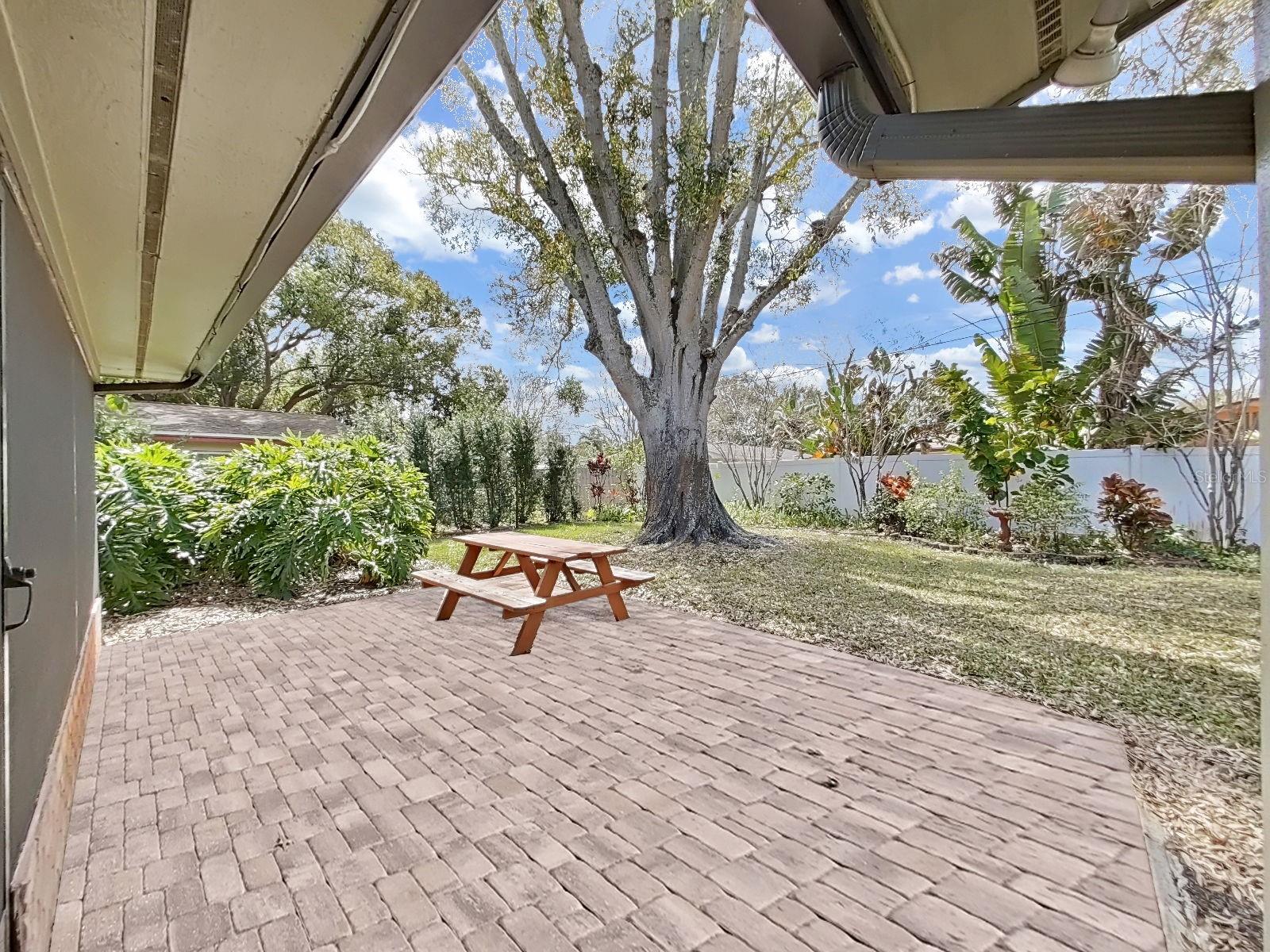 Brick paver patio (with included picnic table) sits beneath a majestic mature shade tree, surrounded by landscape and yard for pets and play- a true backyard oasis!