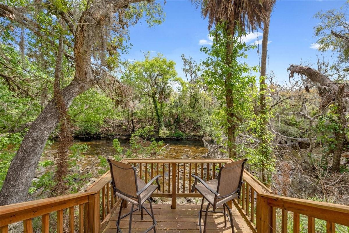 Multi-level wood deck overlooks the Alfia River, framed by mature trees and lush natural landscaping.