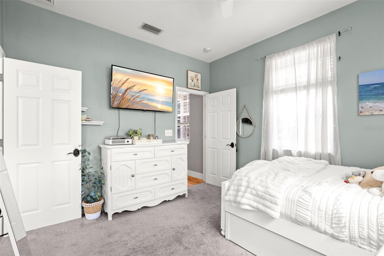 Light-filled second bedroom with plush carpeting, neutral tones, ceiling fan, and a large window.
