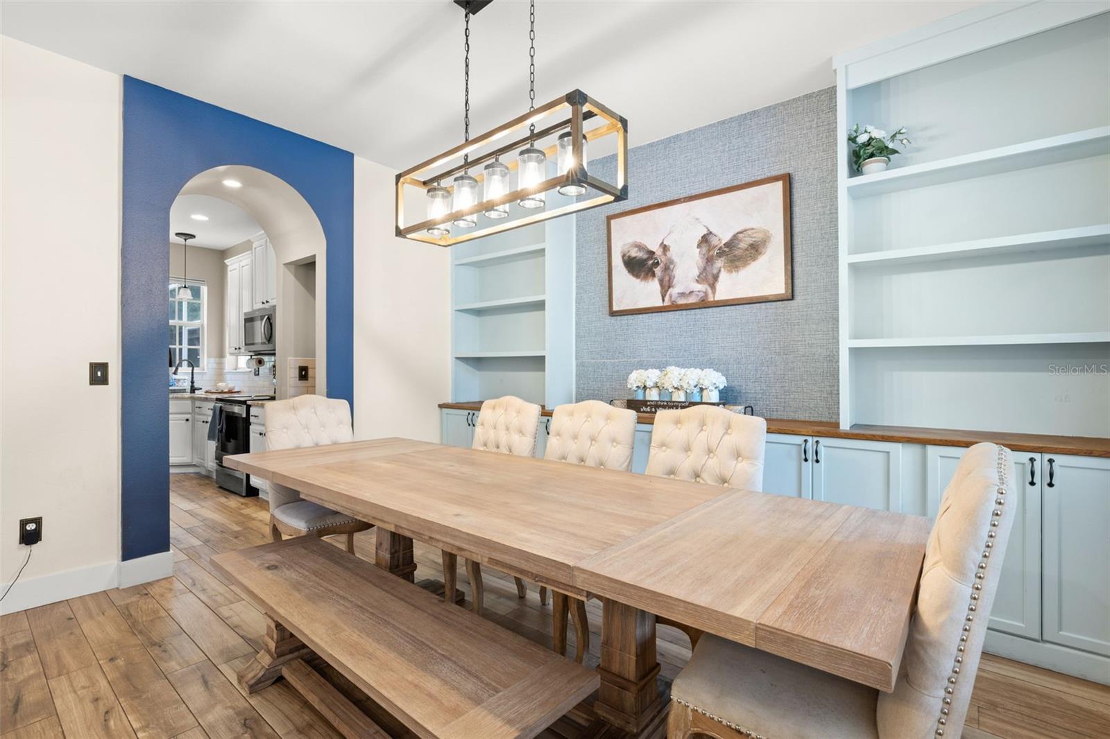 Show-stopping dining room with custom built-in shelving and cabinetry, a striking linear chandelier, and architectural details including a bold accent wall and arched opening to the kitchen.