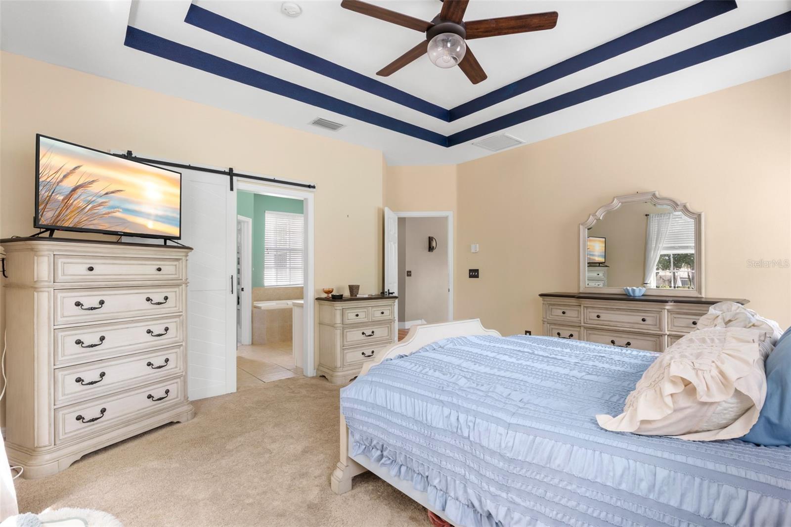 Primary suite with tray ceiling accented by painted trim, ceiling fan, and soft carpeting, complemented by a sliding barn door entry to the ensuite bath and a light, neutral color palette.