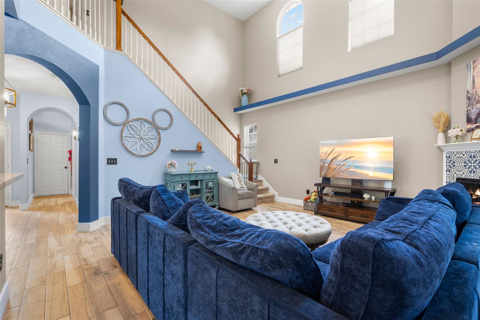 Living room view highlighting soaring ceilings, clerestory windows, and wood plank flooring, with a staircase featuring a wood banister and white spindles. Arched openings and painted accent ledges add architectural character.