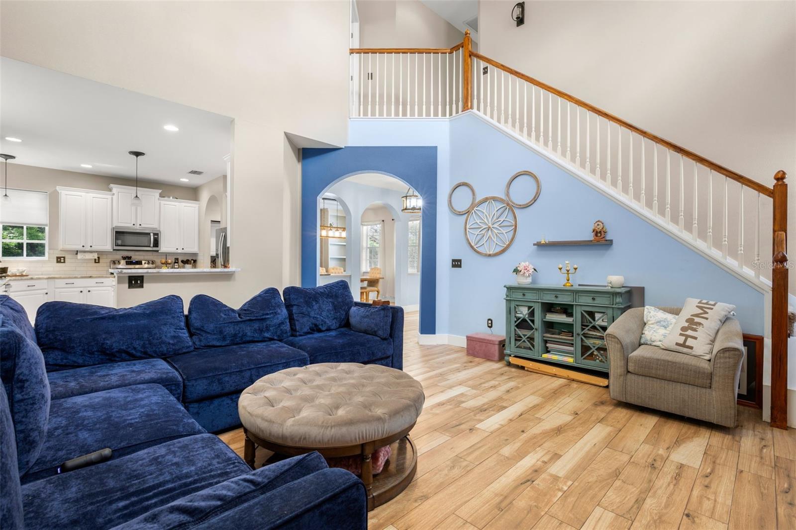 Spacious living area with wood plank flooring and a staircase with wood banister, complemented by a painted accent wall and arched openings that lead to both the dining room and kitchen.