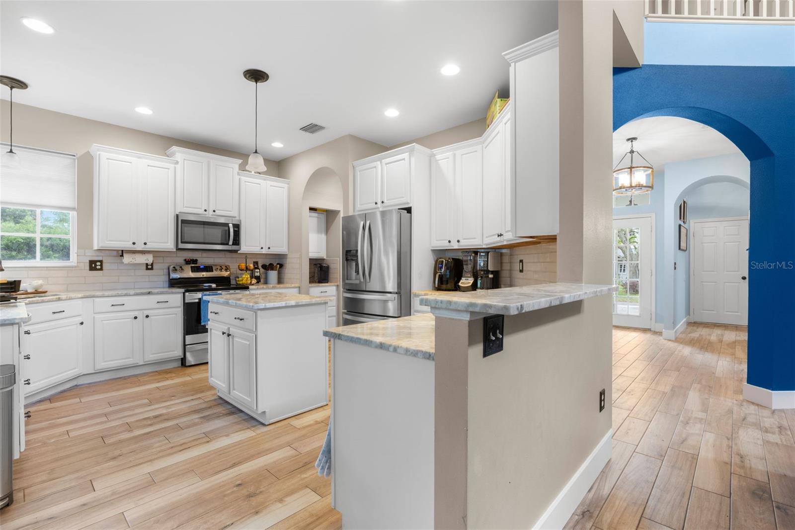 Expansive kitchen with dual islands, marble countertops, stainless steel appliances, and alabaster cabinetry, enhanced by arched openings and continuous wood plank flooring.