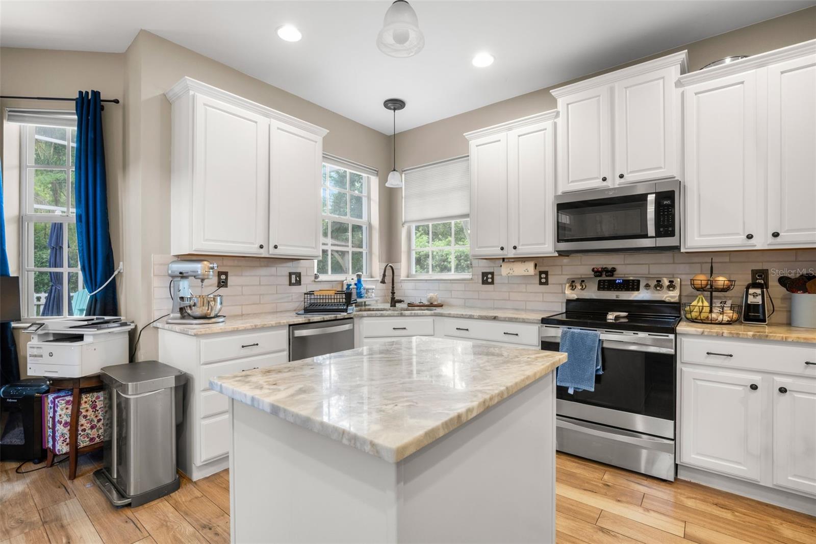 Bright, functional kitchen featuring a central island with marble countertops, a corner sink beneath dual windows, alabaster cabinetry, subway tile backsplash, and stainless steel appliances.