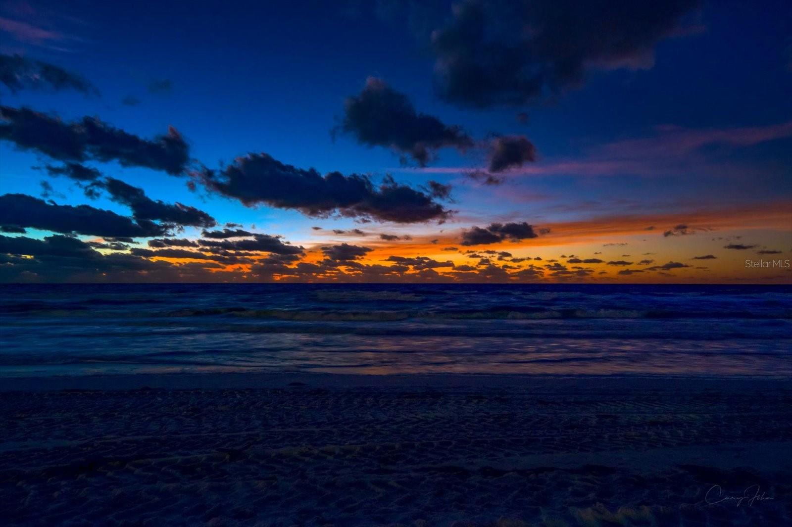 Deep indigo waves meet a vibrant horizon of fiery amber and soft violet as dusk settles over the peaceful shores of Indian Rocks Beach.
