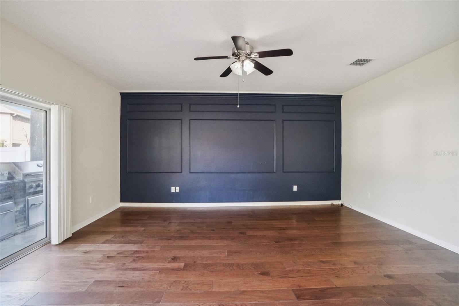 Living Room - The spacious living room makes a bold design statement with a full-wall navy board-and-batten accent wall, rich hardwood flooring, a ceiling fan, and a sliding glass door leading to the outdoor space a stylish and comfortable gathering area.