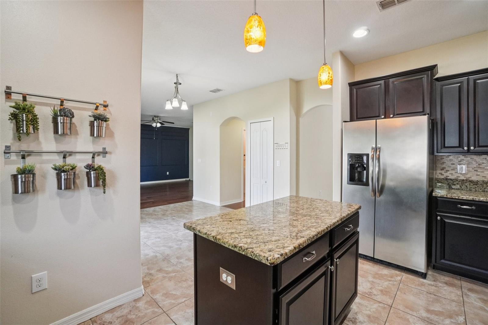 This view from the kitchen island looks toward the open living and dining areas, showcasing the home's connected layout, arched doorways, tile flooring transitioning to wood-look flooring, and a decorative wall-mounted herb planter display.