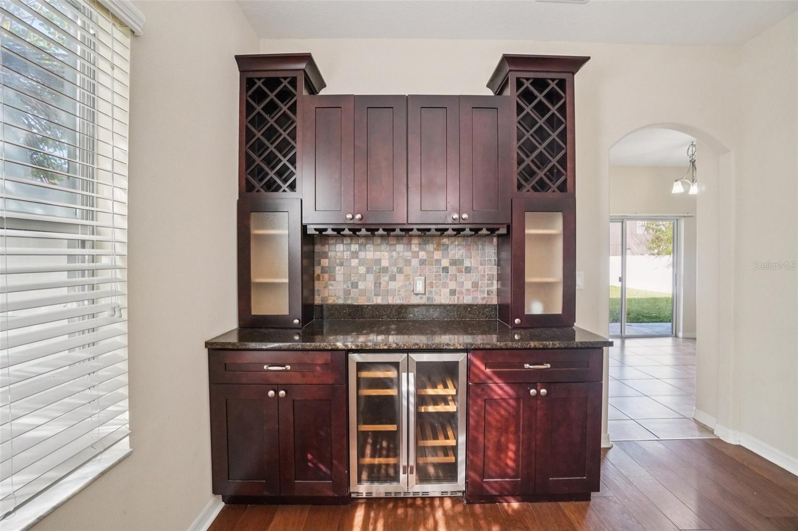 Custom built-in wet bar with dark cherry cabinetry, lattice wine rack, stemware hanging rack, glass-front display cabinets, dark granite countertop, mosaic tile backsplash, and stainless steel wine cooler.