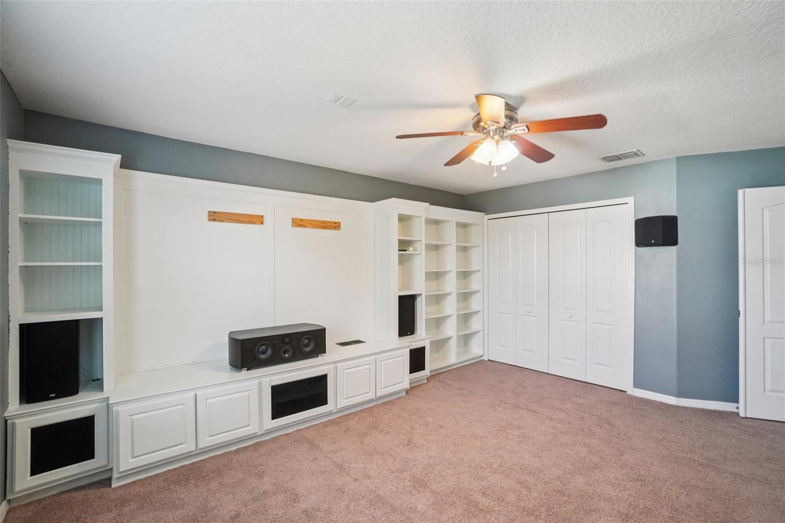 Secondary bedroom with full custom built-in entertainment wall, flanking open bookshelves, large closet, plush carpet, and ceiling fan.