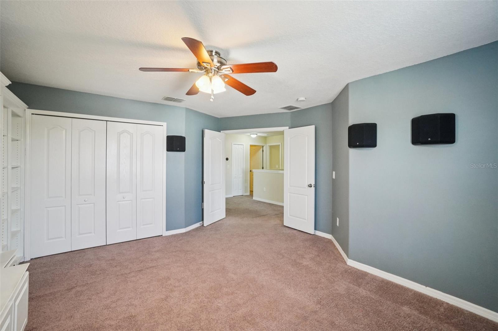 Spacious secondary bedroom with dual closets, wall-mounted surround sound speakers, ceiling fan, plush carpet, and hallway access.