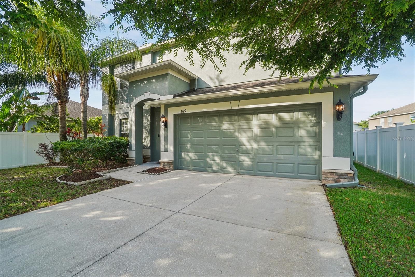 Wide concrete driveway, two-car garage, and manicured landscaping framed by mature shade trees.